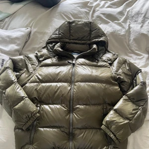 Olivgrön pufferjacka från Polo Ralph Lauren - Snygg olivgrön pufferjacka från Polo Ralph Lauren med broderad logga på bröstet. Jackan har huva, dragkedja framtill och två sidofickor med dragkedja. Perfekt för kalla dagar och ger en cool vibe till din outfit. Observera litet skav på sista bilden