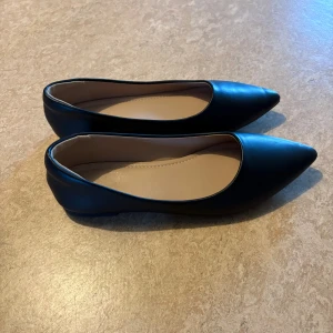 Svarta ballerina skor  - Stilrena svarta ballerinaskor i syntetmaterial med spetsig tå och låg platt sula. Skorna har en klassisk och minimalistisk design som passar till många olika outfits. Insidan är beige och mjukt vadderad för extra komfort.