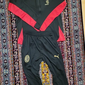 AC Milan x Puma svart träningshoodie - Svart träningshoodie från Puma och AC Milan med röda detaljer längs ärmarna och guldiga loggor. Långärmad modell med half zip och huva, tillverkad i lätt polyester. Perfekt för fotbollsträning eller chill. Snygg och sportig stil.