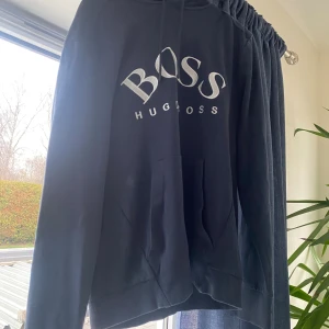 Svart hoodie från Hugo Boss - Svart hoodie från Hugo Boss med stor vit logga på bröstet. Klassisk modell med huva, snörning och känguruficka framtill. Perfekt för en chill och stilren look.