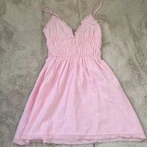 Ljusrosa kort klänning från Bershka - Supersöt kort ljusrosa klänning från Bershka med smala axelband och v-ringning. Klänningen har resår i midjan för en snygg passform och volangdetaljer vid urringningen. Perfekt för varma dagar och somriga vibes! Aldrig använd