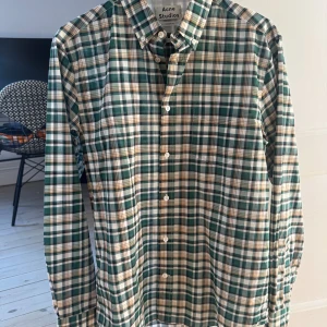 Rutig skjorta från Acne Studios - Snygg rutig skjorta från Acne Studios i grönt, vitt och brunt. Klassisk krage, knäppning framtill och lång ärm. Tillverkad i en mix av bomull och nylon för en lätt, tunn och bekväm känsla. Perfekt för dig som gillar stilrena och trendiga plagg. Aldrig använd.