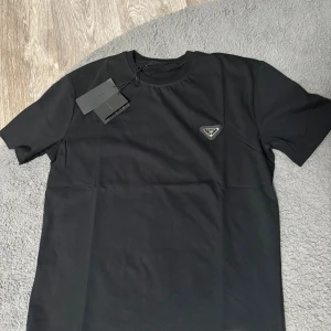 Svart t-shirt från Prada med logga - Snygg svart t-shirt från Prada med klassisk rund hals och korta ärmar. På bröstet sitter en ikonisk triangelformad metallogo. T-shirten är i mjuk bomull och har en clean, stilren look som passar perfekt till streetwear eller en mer dressad stil.