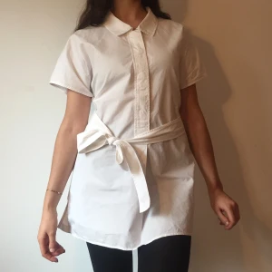 Vit skjortklänning med knytband - Vit kort skjortklänning med klassisk krage och korta ärmar. Klänningen har ett snyggt knytband i midjan. 100% bomull. I storlek M, men modellen på bilden har storlek S. 