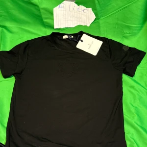 Svart Moncler t-shirt  - Svart t-shirt från Moncler med diskret broderad logga på bröstet och patch på ärmen. Klassisk rund hals och korta ärmar. Tillverkad i mjuk bomull för en skön känsla. Perfekt för en clean och stilren look.Liknande modell som originalet!