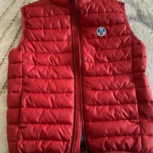 Röd dunväst från North Sails - Röd quiltad dunväst från North Sails med dragkedja framtill och rund hals. Västen har ett broderat North Sails-märke på bröstet och svart foder med stor logga på insidan. Perfekt för lager-på-lager under kyliga dagar.