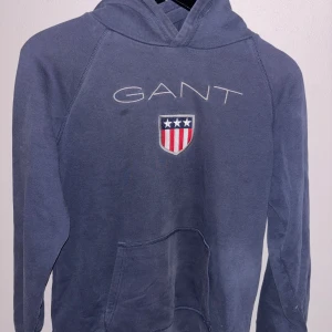 Blå hoodie från Gant med broderad logo - Säljer en blå hoodie från Gant med broderad logotyp och sköld på bröstet. Tröjan har huva, långa ärmar och en stor magficka framtill. Perfekt för dig som gillar klassisk och sportig stil.