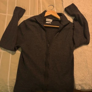 Mörkgrå Merinoulls Full Zip från These Glory Days - Säljer en stilren mörkgrå kofta från These Glory Days i mjuk och exklusiv merinoull. Koftan har hel dragkedja framtill, hög krage och långa ärmar. Perfekt för lager-på-lager och snygg till både jeans och chinos. STORLEK L MEN VÄLDIGT LITEN PASSAR M-S