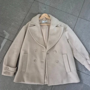 Beige skepparkavaj från Ellos, stl 38 - Stilren beige skepparkavaj från Ellos i klassisk modell med bred krage, dubbelknäppning och två fickor framtill. Perfekt för dig som vill ha en tidlös look.🎀