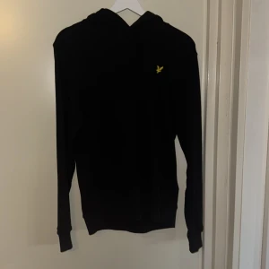 Svart hoodie från Lyle & Scott Storlek S - Svart hoodie från Lyle & Scott med klassisk gul logga på bröstet. Tröjan har huva med snörning och långa ärmar. Tillverkad i mjuk bomull, perfekt för en chill och stilren look. Storlek S och använd 1 gång inga defekter!