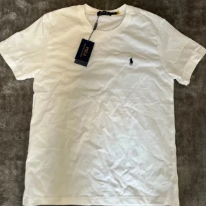 Vit t-shirt från Polo Ralph Lauren - Klassisk vit t-shirt från Polo Ralph Lauren med rund hals och kort ärm. Ikoniska broderade loggan på bröstet. Tillverkad i mjuk bomull för en skön känsla. Perfekt basic-plagg med stilren design.