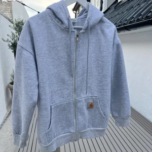 Grå hoodie med dragkedja från Carhartt - Snygg ljusgrå hoodie från Carhartt med dragkedja framtill, huva med snörning och två stora fickor. Klassisk logga på vänster ficka. Perfekt för chill dagar och streetwear-stil. Mjuk bomullskänsla och relaxed fit.