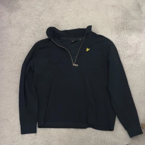 Svart half zip tröja Lyle & Scott - Svart långärmad tröja från Lyle & Scott med half zip och klassisk gul logga på bröstfickan. Tröjan har en enkel och stilren design med mjukt material och ribbade muddar. Perfekt för lager-på-lager och chill vardagsstil.