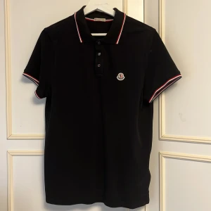 Moncler piké - Svart pikétröja från Moncler med klassisk krage och knäppning framtill. Tröjan har röda och vita ränder på krage och ärmslut samt Moncler-logga på bröstet. Perfekt för en stilren och sportig look.