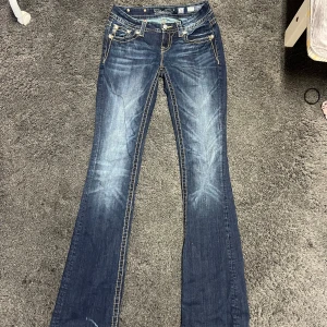 Miss Me bootcut jeans med paljettdetaljer - Snygga blå bootcut jeans från Miss Me med coola paljettdetaljer och broderier på bakfickorna. Jeansen har kontrastsömmar, midwaist passform och klassisk femficksdesign. Perfekta för dig som vill sticka ut med extra detaljer och unik stil. Strl 26 men känns mer som en 28 men innerbenslängden 36 inches/ 92 cm så passar bra till tjejer med långa ben.