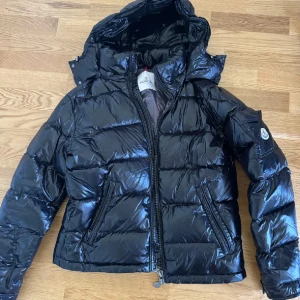 Svart pufferjacka från Moncler - Snygg svart pufferjacka från Moncler med glansig finish och huva. Jackan har dragkedja framtill, två fickor med dragkedja och klassisk Moncler-logga på ärmen. Perfekt för kalla dagar när du vill hålla stilen.