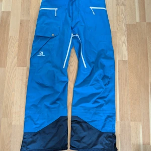 Blå täckbyxor från Salomon - Blå täckbyxor från Salomon med vita dragkedjor, flera fickor och förstärkta svarta partier vid bensluten. Perfekta för vinterns äventyr och har en sportig look. Materialet är slitstarkt syntet och byxorna har en loose passform. Ventilation i grenen. 