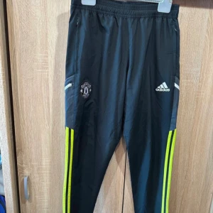 Manchester United Adidas träningsbyxor M - Svarta träningsbyxor från Adidas med Manchester United-logga på benet och vita Adidas-detaljer. Byxorna har neongula ränder längs sidorna, dragkedjor vid fickorna och elastisk midja. Perfekta för fotboll eller träning.