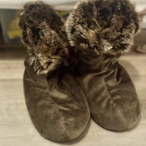 Bruna inneskor med fuskpäls - Mysiga bruna inneboots med mjuk fuskpäls upptill och mockaliknande material på utsidan. Perfekta för kalla dagar när du vill hålla fötterna varma och snygga. De har en rund tå och platt sula för extra komfort. Perfekt inför höst/vinter.