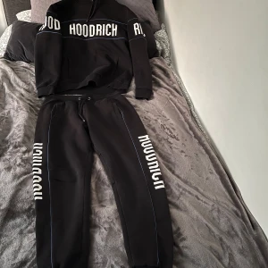 Svarta mjukisbyxor och hoodie från Hoodrich  - Svarta mjukisbyxor och hoodie från Hoodrich med vit logga längs benet och tunna blå detaljer. Byxorna har snörning i midjan och ribbade muddar vid benslut. Perfekta för en chill och street-inspirerad look.