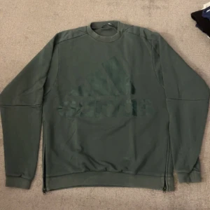 Mörkgrön Adidas sweatshirt med dragkedjor - Snygg mörkgrön sweatshirt från Adidas med stort ton-i-ton logotryck på bröstet. Tröjan har långa ärmar, ribbade muddar och coola dragkedjor i sidorna för extra stilpoäng. Perfekt för dig som gillar sportig och avslappnad look. Oversized tröja 