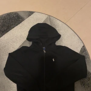 Svart hoodie från Polo Ralph Lauren XS - Svart hoodie från Polo Ralph Lauren i 100% bomull. Klassisk design med dragkedja framtill, huva och den ikoniska broderade loggan på bröstet. Perfekt för en chill och stilren look.