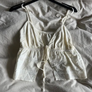 Vit linnetopp med knytband - Söt vit linnetopp med tunna axelband och knytning framtill. Toppen har volangliknande lager och dekorativa sömmar som ger en bohemisk känsla. Perfekt för varma dagar och enkel att styla med jeans eller kjol.