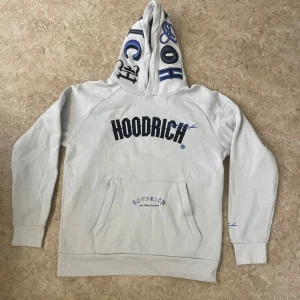 Vit hoodie från Hoodrich med tryck - Snygg vit hoodie från Hoodrich med svart och blått tryck på bröstet och huvan. Stora broderade bokstäver på huvan och textdetaljer på ärm och rygg. Klassisk känguruficka framtill och ribbade muddar. Perfekt för en streetwear-look.