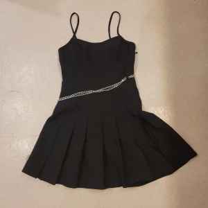Svart kort klänning med kedjedetalj - Svart kort klänning med tunna axelband och utställd kjol med veck. Snygg silverfärgad kedja i midjan som ger en edgy touch. Perfekt för dig som gillar minimalistisk stil med en twist.
