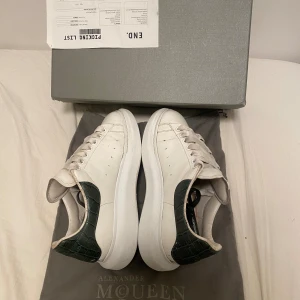 Alexander McQueen vita sneakers - Säljer ett par stilrena vita sneakers från Alexander McQueen med chunky plattformssula och gröna detaljer i croco-mönstrad skinn på hälen. Klassisk rund tå och snörning framtill. Perfekta för dig som vill ha en clean look med lyxig touch.