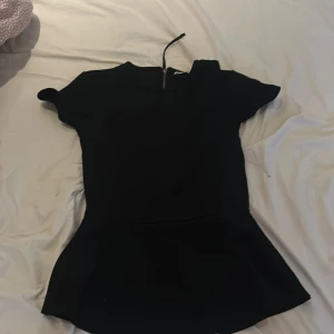 Svart kort topp från C, XS - Supersnygg svart pepplum topp från C i storlek XS. Toppen har korta ärmar, rund halsringning och markerad midja med utställd kjol. Dragkedja i ryggen ger en cool detalj. Perfekt för dig som gillar stilrena och enkla plagg.