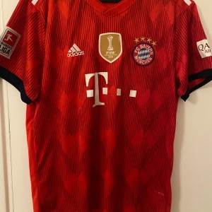 Lewandowski 19/20 Bayern München tröja - Säljer en FC Bayern München matchtröja från Adidas med Lewandowski och nummer 9 på ryggen i storlek M. Tröjan är röd med subtilt mönster, vita tryck och svarta detaljer vid ärmslut. Klassisk V-ringning, kortärmad och tillverkad i lätt funktionsmaterial. Bundesliga och FIFA-märken på ärmar och bröst. Har lite slitage på trycket där fram och på nummer 9