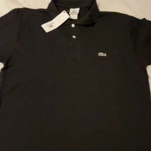 Svart pikétröja från Lacoste - Klassisk svart pikétröja från Lacoste med vit knappslå och ikoniska krokodilloggan på bröstet. Tröjan har krage och korta ärmar, perfekt för en clean och sportig stil. Tillverkad i mjuk bomull som andas.