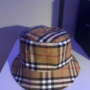 Burberry bucket hat  - Säljer en vändbar bucket hat från Burberry. Ena sidan har klassiskt beige, svart, rött och vitt rutmönster, andra sidan är helsvart med vit Burberry-logga framtill. Hatten är i bomull och har en avslappnad passform. Perfekt för dig som vill ha två stilar i en.