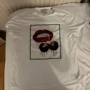 Vit Limitato t-shirt med körsbärstryck - Vit t-shirt från Limitato med coolt tryck av röda läppar och två körsbär på bröstet, designad av Sara Pope. Klassisk passform och rund hals. Perfekt statement-plagg för dig som vill sticka ut.