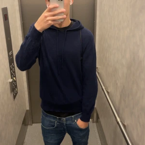 Mörkblå hoodie med huva - Mörkblå hoodie med huva och snörning. Tröjan har en avslappnad passform och är långärmad, perfekt för kyligare dagar. Enkel och stilren design som funkar till det mesta.