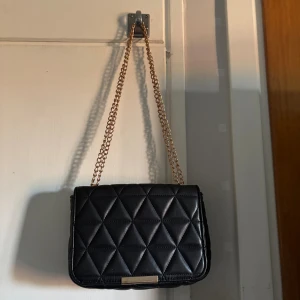 Svart axelväska med kedja - Snygg svart axelväska med quiltat mönster och guldfärgad kedjerem. Väskan har en klassisk rektangulär form, magnetlås och innerfack med dragkedja. går att ha kedjan på två olika sätt som bilderna visar. Tror den är ifrån Glitter 