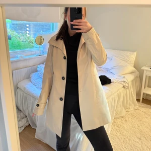Beige trenchcoat från Bondelid - Stilren beige trenchcoat från Bondelid med klassisk krage och svarta knappar. Jackan har långa ärmar och snygga detaljer vid axlarna. Storlek Xs