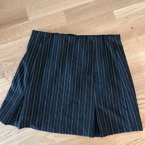 Svart kortkjol med vita ränder - Snygg svart kortkjol med smala vita vertikala ränder. Kjolen har en rak passform och är tillverkad i ett mjukt tyg som känns bekvämt mot huden. Perfekt för dig som gillar en stilren och modern look med lite edge.