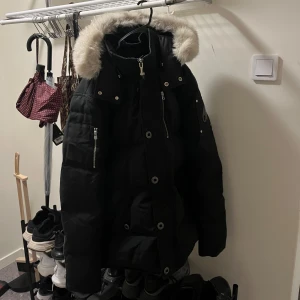 Svart dunjacka Moose Knuckles med pälskrage - Säljer en svart dunjacka från Moose Knuckles med fluffig pälskant på huvan. Jackan har dragkedja, knappar och flera fickor med dragkedja. Perfekt för kalla dagar och har en riktigt snygg, sportig look.