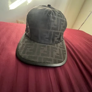 Svart Fendi keps med logomönster - Säljer en svart keps från Fendi med ikoniskt FF-logomönster över hela kepsen. Kepsen har böjd skärm med svart kant och justerbar rem baktill med Fendi-märke. Materialet är textil med exklusiv känsla och kepsen har en stilren, urban look.