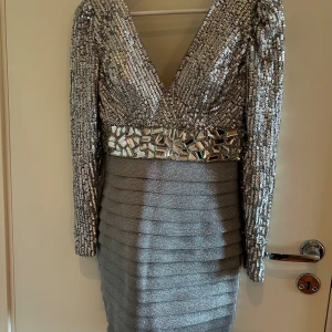 Glittrig silverklänning från Mac Duggal - Superglittrig silverfärgad kortklänning från Mac Duggal med paljetter och spegelstenar upptill, markerad midja och djup v-ringning. Långa ärmar och figurnära passform i skimrande polyester. Perfekt för dig som vill sticka ut och glänsa på festen!