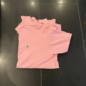 Rosa pikétröja Polo Ralph Lauren XL - Säljer en klassisk rosa pikétröja från Polo Ralph Lauren i storlek XL. Tröjan har korta ärmar, krage med knappar och den ikoniska gröna loggan broderad på bröstet. Tillverkad i 100% bomull för en skön och luftig känsla.