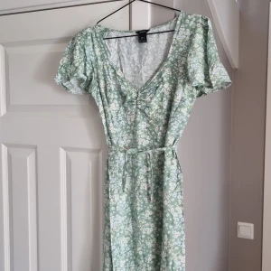 Grön blommig långklänning Lindex - Supersöt långklänning från Lindex i ljusgrönt med små vita och ljusblå blommor. Klänningen har v-ringning med rynkdetalj, korta puffärmar och ett knytband i midjan. Perfekt för sommarens alla tillfällen!