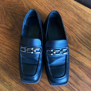 Svarta loafers med metallkedja - Stilrena svarta loafers i skinn med fyrkantig tå och chunky låg klack. Snygg detalj med silverfärgad kedja över vristen som ger en edgy vibe. Perfekta för dig som gillar klassiska skor med en twist. Fint skick, använd ca 1-2 gånger. Storlek 39, men lite små i storleken då de är smala. Säljer för 200 kr