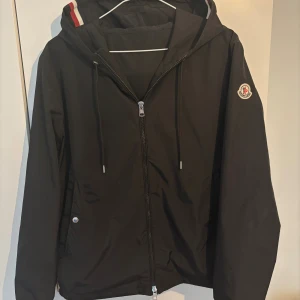 Svart vindjacka från Moncler med huva - Svart vindjacka från Moncler med huva och dragkedja framtill. Jackan har Moncler-logga på ärmen, snörning i huvan och diskreta fickor med knapp. Snygg detalj med röd och vit rand på huvan. Perfekt för dig som vill ha en stilren och exklusiv look.