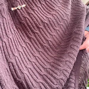 Brun stickad poncho med fransar - Säljer en brun stickad poncho med snyggt kabelstickat mönster och fransar längs kanten. Perfekt att slänga över en hoodie eller tröja för extra värme. Fäst med en dekorativ brosch framtill. Supermysig för kyliga dagar!