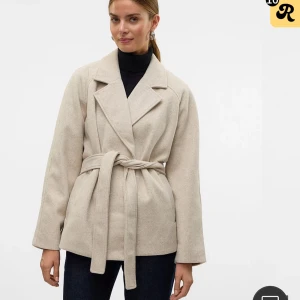 Beige kappa med bälte från Vero Moda - Stilren beige kappa från Vero Moda i storlek M. Jackan har klassisk krage, knytskärp i midjan och långa ärmar. Materialet är mjukt och känns ulligt, perfekt för kyligare dagar. Enkel och clean look som passar till det mesta. fickan har lossnat lite men syns knappt och kan sys ihop.