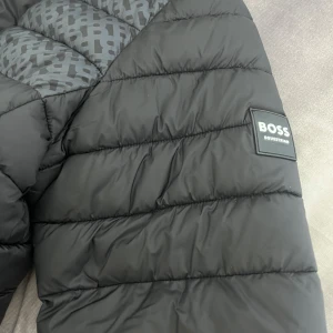 Svart pufferjacka från BOSS Equestria - Säljer en svart puffer vinterjacka från BOSS Equestrian med huva och dragkedja. Jackan har mönstrade partier med BOSS-logga, två dragkedjefickor och snygga ränder i vitt och beige på huvan. Perfekt för kalla dagar och riktigt stilren.