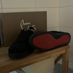 Christian Louboutin svarta sneakers med nitar - Svarta sneakers från Christian Louboutin med ikoniska röda sulor och svarta nitar över tån. Skorna har snörning och en chunky platt sula. Tillverkade i skinn och textil, perfekt för dig som vill sticka ut med en edgy och lyxig stil.